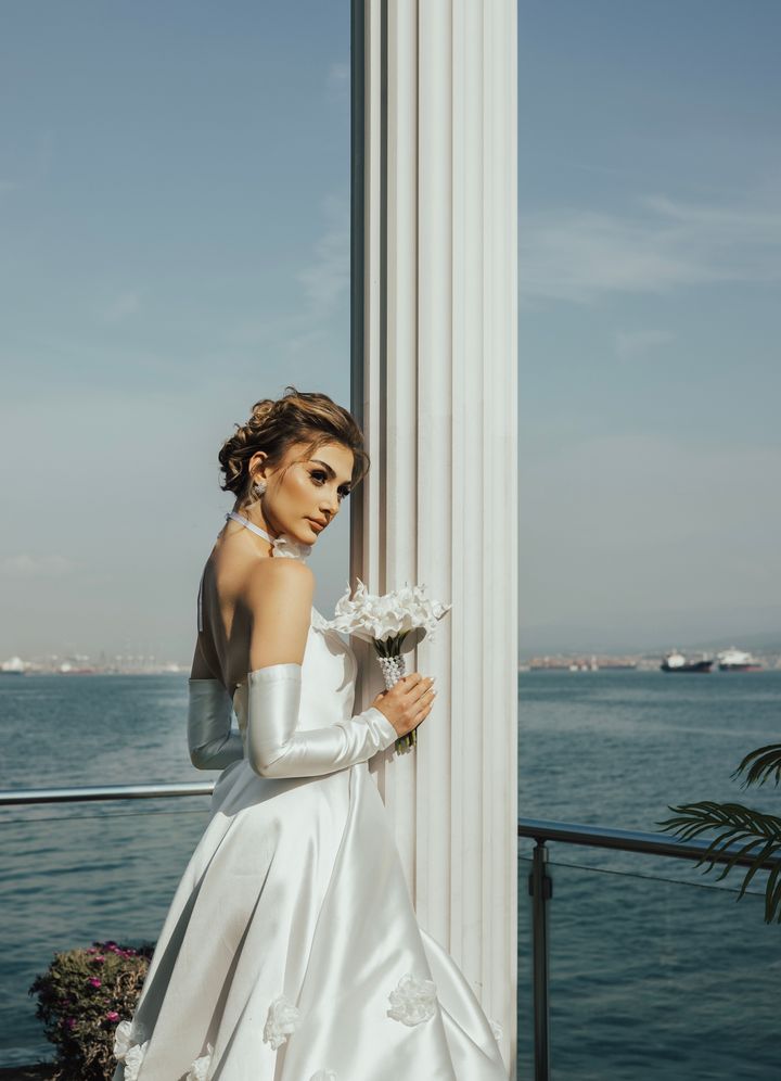 A bride in a white gown standing by a window overlooking the sea