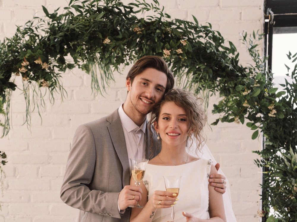 A smiling couple holding champagne glasses beneath a greenery arch