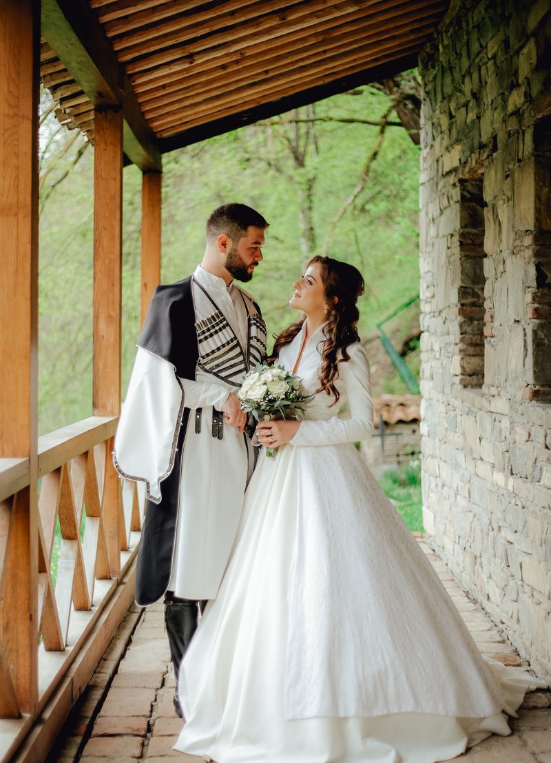 A bride in a ball gown and a groom in traditional Georgian dress on a wooden balcony