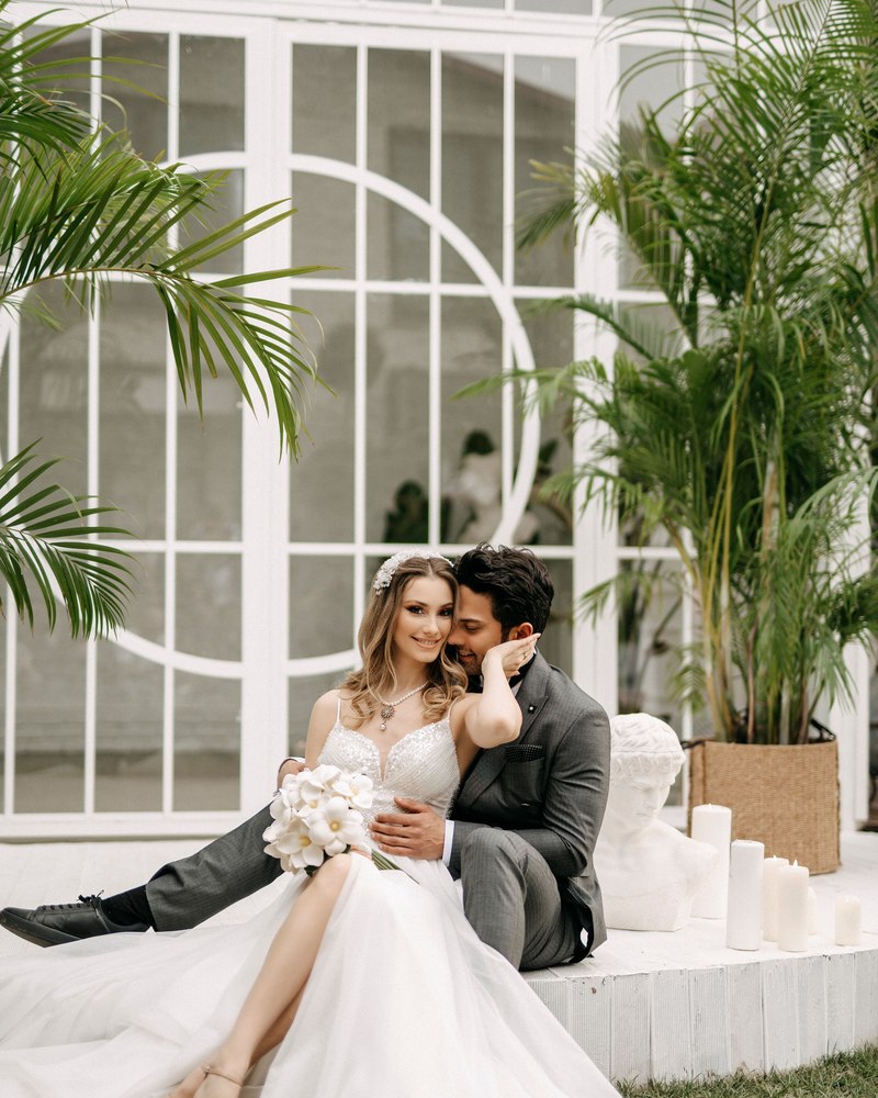 A bride and groom sitting together on steps inside a glass and palm-filled venue