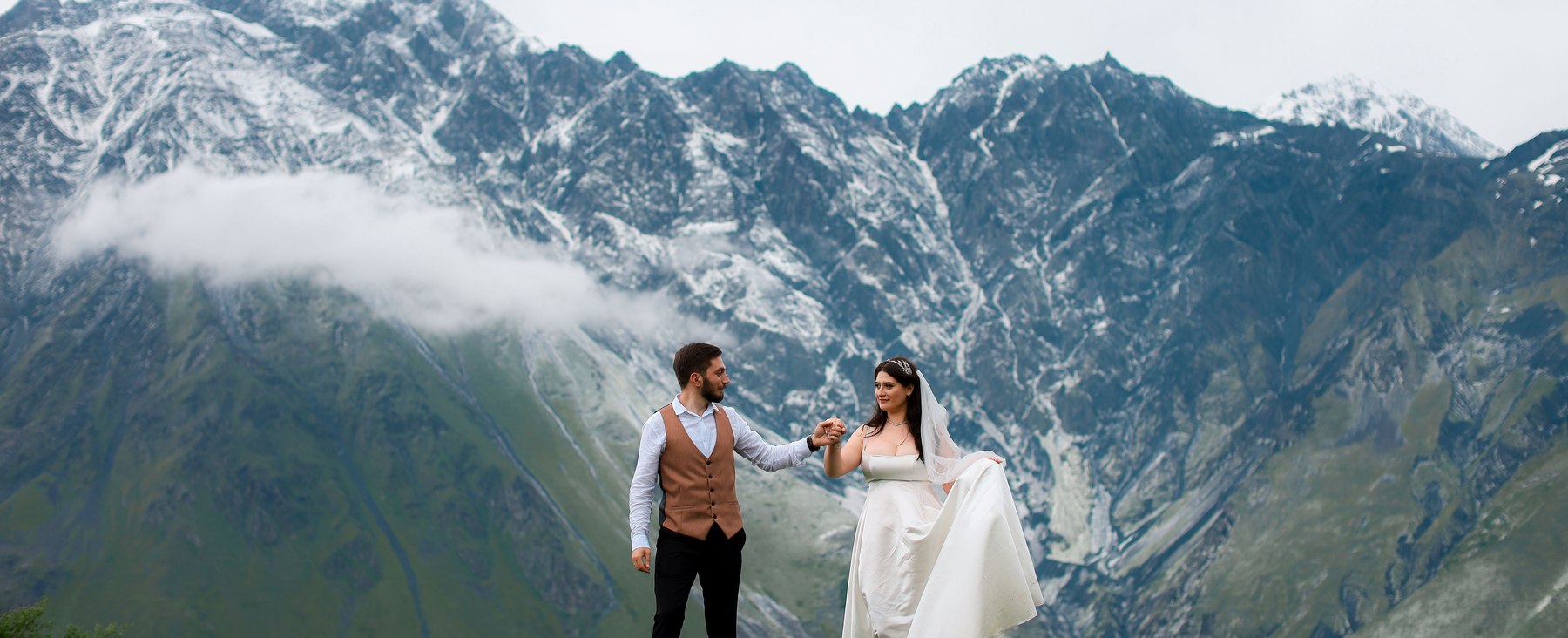 A bride and groom holding hands against the dramatic snow-capped Caucasus Mountains in Kazbegi