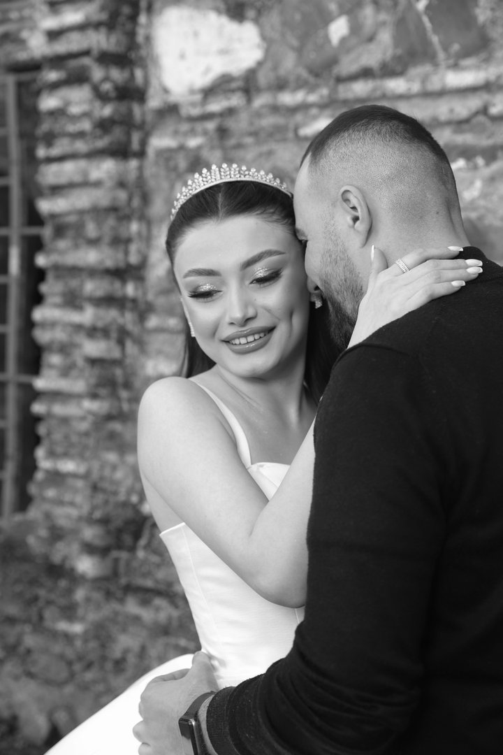 A bride wearing a tiara embraces her groom in front of an old stone wall