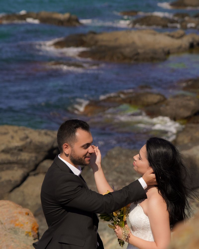 A groom gently touches his bride's face on rocks by the sea