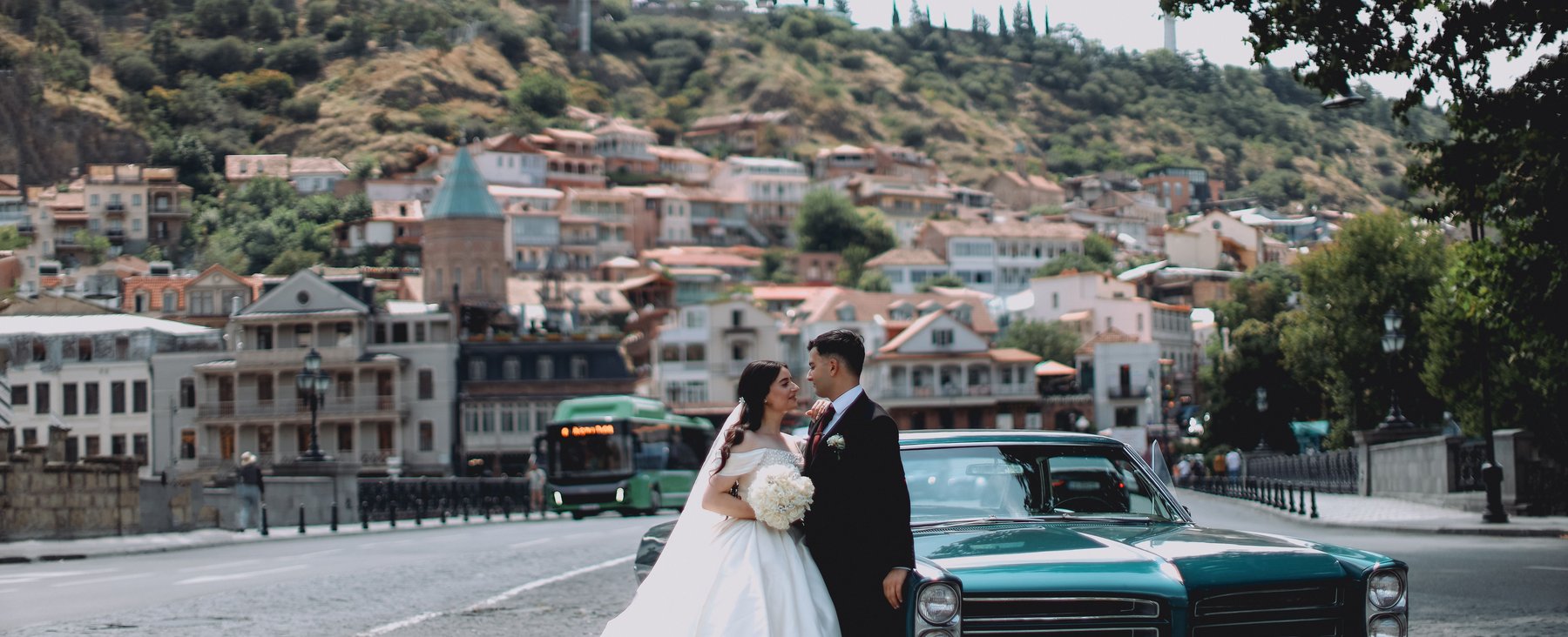A bride and groom pose beside a vintage car with the Tbilisi old town and Narikala fortress behind them
