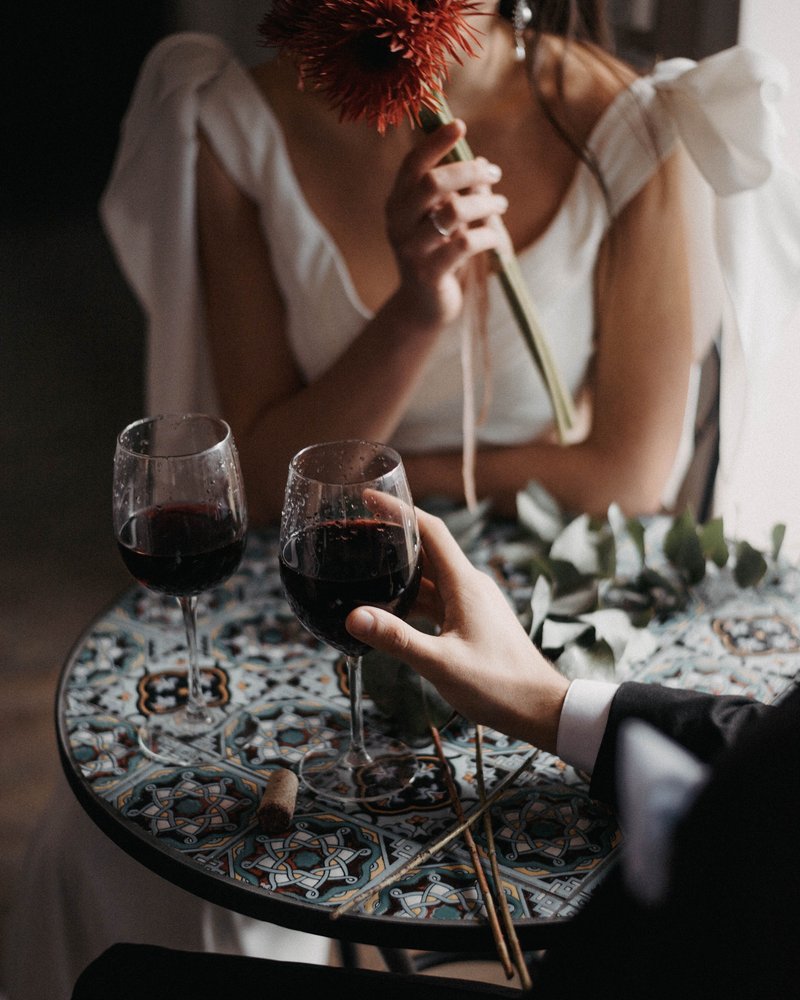 A bride holding a flower and a groom's hand reaching for wine glasses on a mosaic table