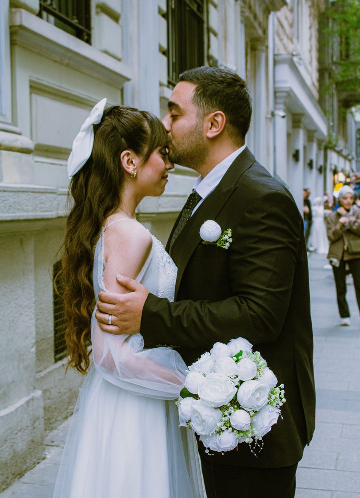 A groom kissing his bride on the forehead on a Tbilisi street, her bouquet of white roses in hand