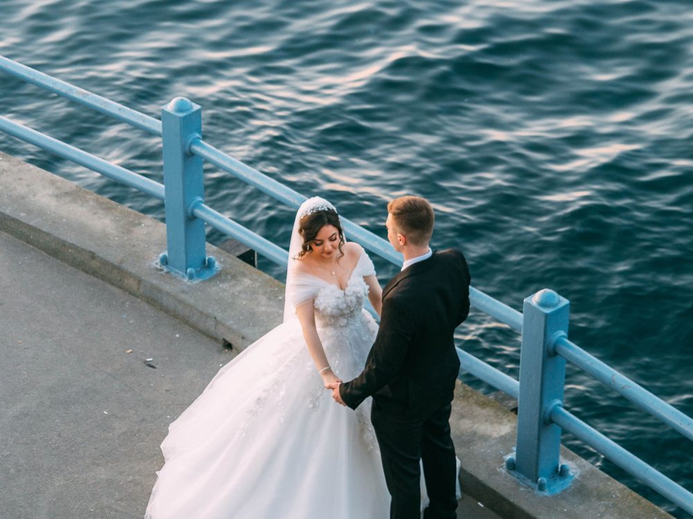 A bride and groom holding hands on a seaside promenade beside a blue railing